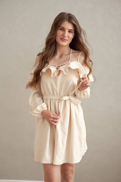 Beautiful young smiling woman portrait with wavy hair, dressed in a beige dress posing in the interior  - Photo, image