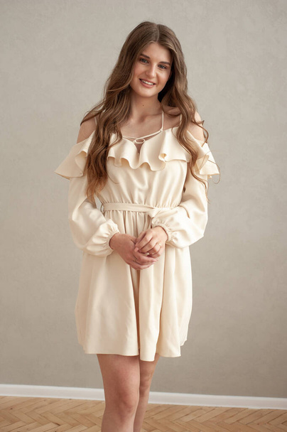 Beau portrait de jeune femme souriante aux cheveux ondulés, vêtue d'une robe beige posant à l'intérieur  - Photo, image