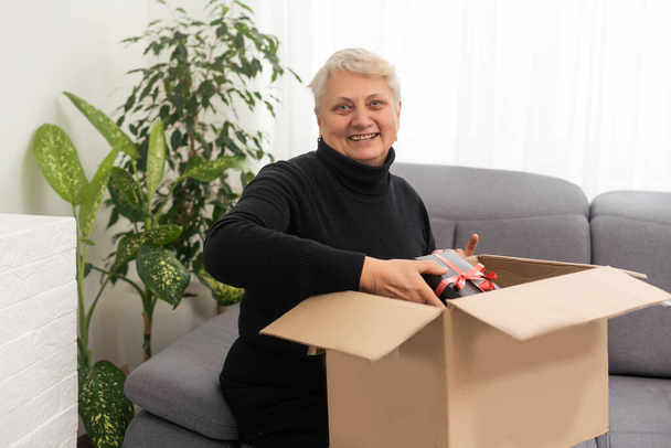 Smiling elderly woman customer receive post shipment parcel at home, happy old senior grandma hold open cardboard box sit on sofa in living room, online shopping order fast courier delivery concept - Foto, Imagen