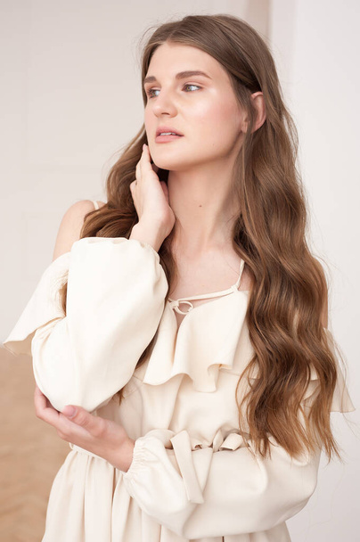 Красивая молодая улыбающаяся женщина портрет с волнистыми волосами, одетый в бежевое платье позирует в интерьере  - Фото, изображение