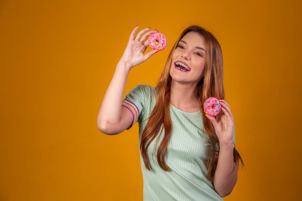 Menina de cabelos vermelhos no fundo amarelo comendo deliciosos donuts coloridos. - Foto, Imagem