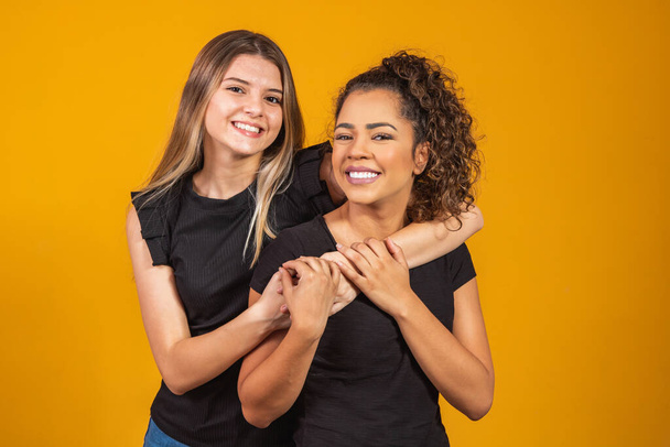 friends of different ethnicities holding each other on yellow background, diversity concept. An afro woman and a blonde hugging each other smiling looking at the camera - 写真・画像