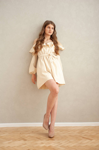 Beautiful young smiling woman portrait with wavy hair, dressed in a beige dress posing in the interior  - Photo, Image