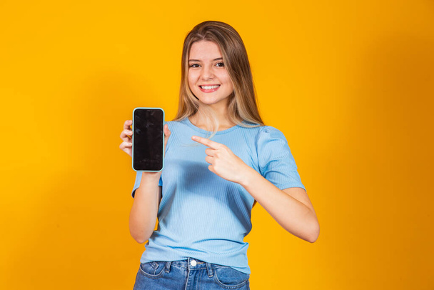 Young blonde girl showing blank smartphone screen and pointing on it while looking at camera and smiling. - Foto, imagen