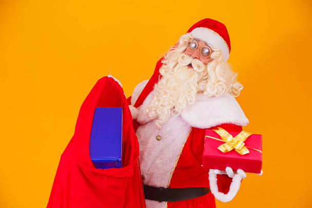 Real Santa Claus on yellow background holding bag with various christmas gifts. - Photo, Image