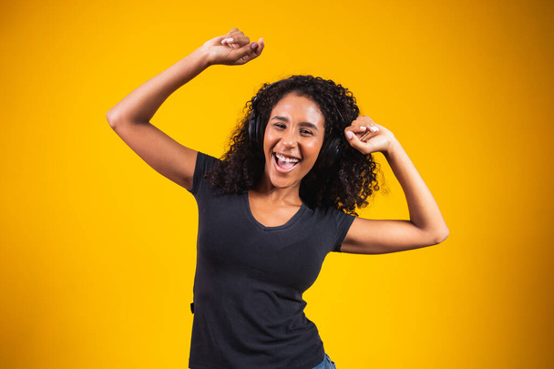 若いアフリカの女性は、音楽を聞いて黄色の壁に分離されたアクティブ踊るヘッドフォンを身に着けている陽気に笑う. - 写真・画像