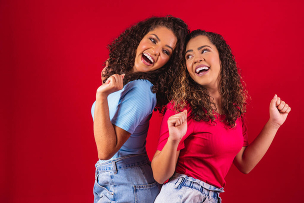 Portrait of happy young afro women friends dancing on red background. - 写真・画像