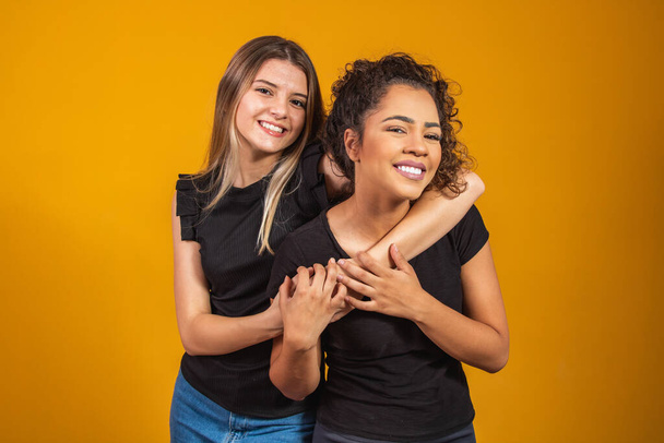 friends of different ethnicities holding each other on yellow background, diversity concept. An afro woman and a blonde hugging each other smiling looking at the camera - Foto, imagen
