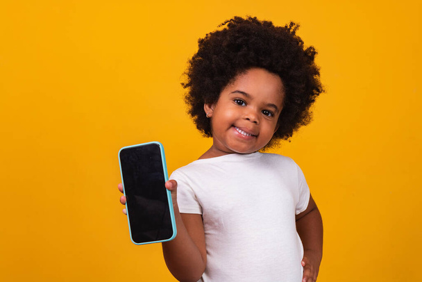 small afro child girl holding a phone and pointing isolated on yellow background. - Valokuva, kuva