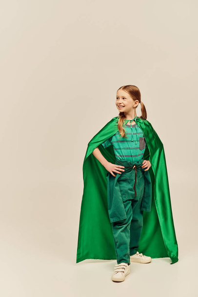 smiling girl in green superhero costume with cloak wearing pants and t-shirt and standing with hands on hips while celebrating International Children Day on grey background  - Foto, imagen