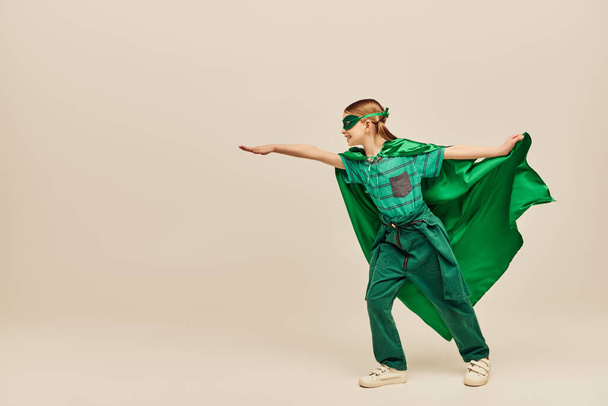 side view of happy kid in superhero costume and mask holding green cloak and standing with outstretched hand while celebrating Child protection day holiday on grey background  - Fotografie, Obrázek