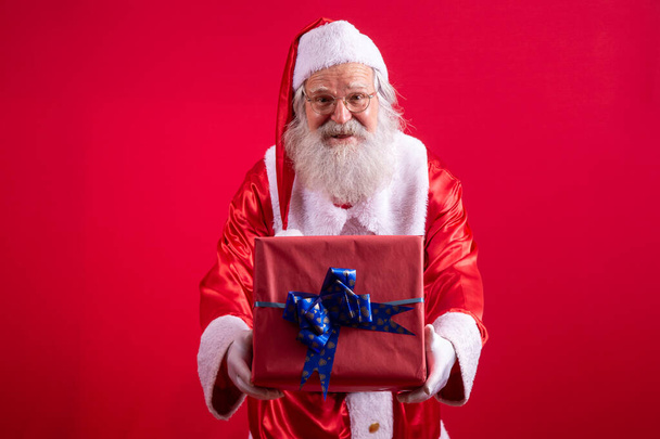 Santa Claus holding a Christmas present on red background. - Fotoğraf, Görsel