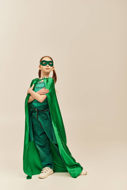 poważna dziewczyna w zielonym stroju superbohatera z płaszczem i maską na twarzy stojącej ze złożonymi ramionami i patrząc w kamerę podczas świętowania wakacji z okazji Dnia Ochrony Dziecka na szarym tle  - Zdjęcie, obraz