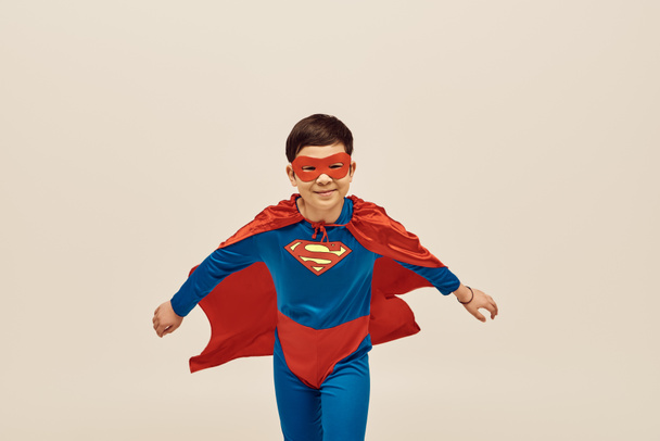 felice ragazzo asiatico in costume da supereroe rosso e blu con mantello e maschera sul viso sorridente mentre celebra la Giornata internazionale per la protezione dei bambini su sfondo grigio  - Foto, immagini