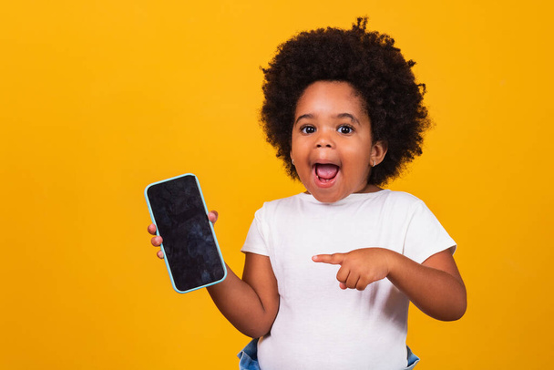 small afro child girl holding a phone and pointing isolated on yellow background. - Photo, image