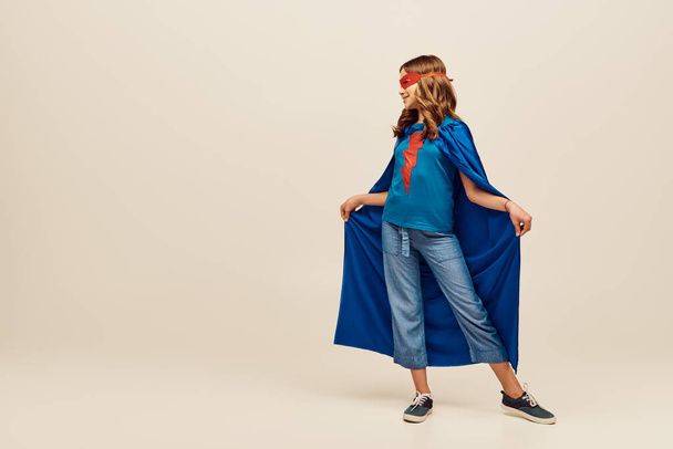 niña feliz en traje de superhéroe y máscara roja en la cara sosteniendo capa azul, de pie en vaqueros y camiseta sobre fondo gris, Día Internacional para la Protección de los Niños concepto  - Foto, imagen