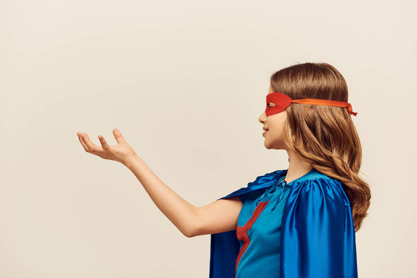 vue latérale de fille heureuse en costume de super-héros avec manteau bleu et masque rouge sur le visage, debout avec la main tendue pendant sur fond gris en studio, concept de Journée mondiale de la protection de l'enfance  - Photo, image