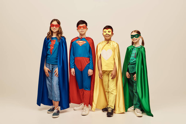fröhliche interrassische Preteen Kids in bunten Superheldenkostümen mit Mänteln und Masken stehen zusammen, während sie den Kinderschutztag auf grauem Hintergrund im Studio feiern  - Foto, Bild