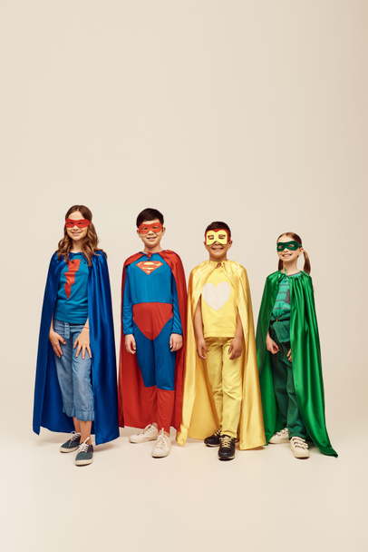 szczęśliwy wieloetniczny nastolatek dzieci w kolorowe kostiumy superbohatera z płaszczami i maskami stojących razem podczas świętowania dzień ochrony dzieci wakacje na szarym tle w studio  - Zdjęcie, obraz