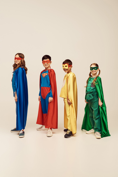 веселые межрасовые дети в разноцветных костюмах супергероев в плащах и масках, стоящих вместе и смотрящих в камеру во время празднования Дня защиты детей на сером фоне в студии  - Фото, изображение