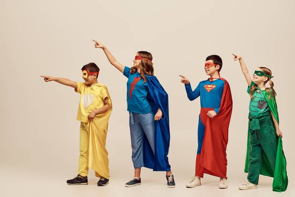 positive multicultural kids in colorful superhero costumes with cloaks and masks pointing with fingers while looking away on grey background in studio, International Day for Protection of Children - Photo, Image