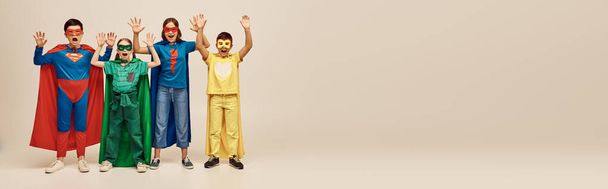 valientes niños interracial en trajes coloridos con capas y máscaras gritando y levantando las manos juntas sobre fondo gris en el estudio, concepto del Día de la Protección del Niño, pancarta  - Foto, imagen