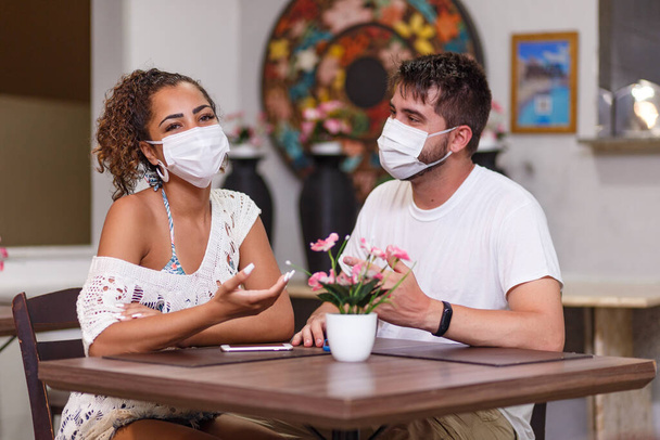 Couple of friends safety talking sitting on an outdoors restaurant wearing safety mask against the covid-19 coronavirus pandemic. New normal life concept - Photo, image