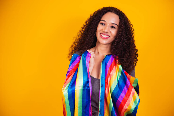 Young curly hair woman covering with lgbt pride flag. Alone. One. Keeping fist up, covering LGBT flag. LGBT+ flag on yellow background. - Φωτογραφία, εικόνα