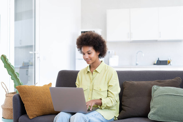 Uśmiechnięta młoda czarna kobieta korzystająca z laptopa, odpoczywająca na wygodnej kanapie, rozmawiająca na rozmowie wideo, czatująca online, przeglądająca internet, oglądająca filmy, robiąca zakupy w Internecie. Leniwy użytkownik gadżetu cyfrowego - Zdjęcie, obraz