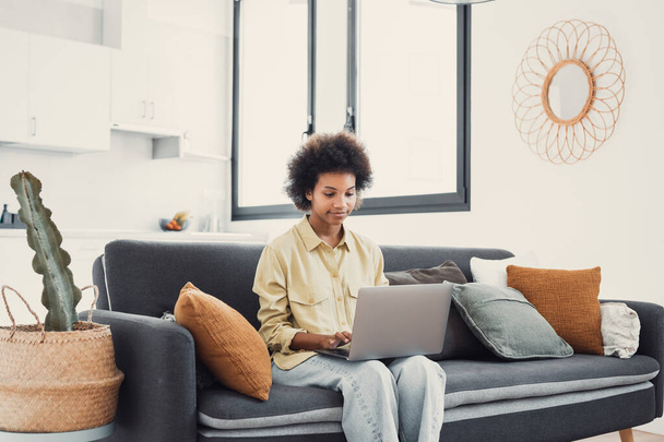 Улыбающаяся молодая чернокожая женщина, пользующаяся ноутбуком, отдыхающая на удобном диване, разговаривающая по видеосвязи, чатящаяся онлайн, просматривающая интернет, смотрящая кино, делающая покупки в интернете. Ленивый пользователь цифрового гаджета - Фото, изображение