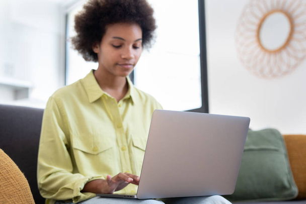 Uśmiechnięta młoda czarna kobieta korzystająca z laptopa, odpoczywająca na wygodnej kanapie, rozmawiająca na rozmowie wideo, czatująca online, przeglądająca internet, oglądająca filmy, robiąca zakupy w Internecie. Leniwy użytkownik gadżetu cyfrowego - Zdjęcie, obraz