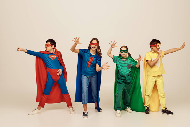 niños multiculturales en trajes de superhéroes coloridos con capas de pie y gestos juntos sobre fondo gris en el estudio, concepto del día internacional de los niños - Foto, imagen