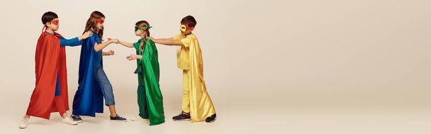 boční pohled na multikulturní děti v pestrobarevných kostýmech superhrdiny s maskami a plášti bojující jeden s druhým na šedém pozadí ve studiu, Mezinárodní dětský koncept, banner - Fotografie, Obrázek