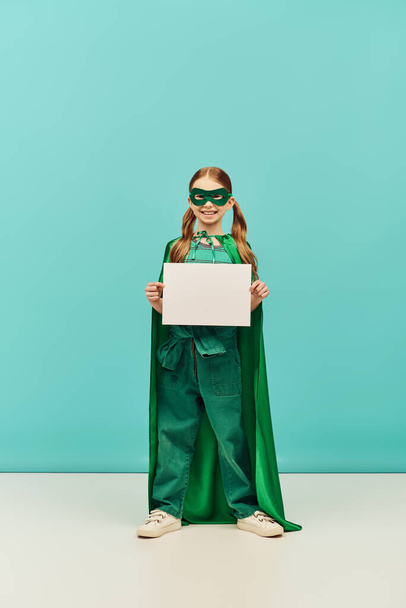 joyeuse fille en costume de super-héros vert avec manteau et masque debout avec du papier vierge et regardant la caméra tout en célébrant les vacances de la journée de protection de l'enfance sur fond bleu  - Photo, image