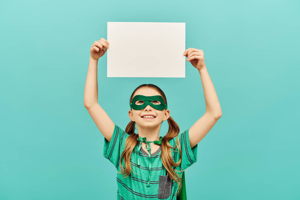 happy preteen girl in green superhero mask holding blank paper above head and looking up on blue background, World child protection day concept  - Foto, Bild