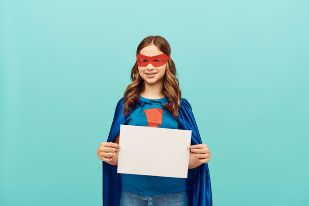 positive preteen girl in superhero costume with red mask standing with blank paper and looking at camera on blue background, World child protection day concept  - Photo, image