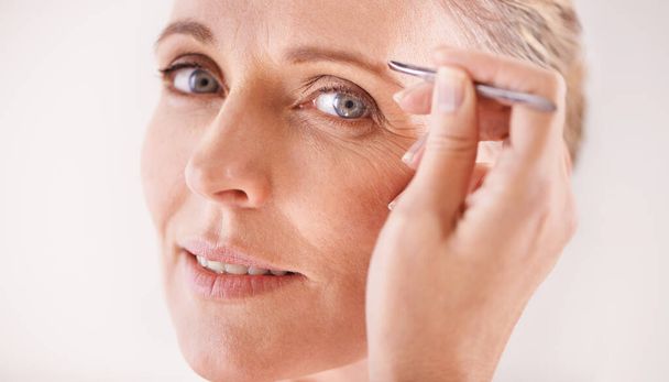 Perfect brows. Portrait of a beautiful mature woman plucking her eyebrows with a pair of tweezers - 写真・画像