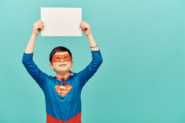 Frühchen asiatischer Junge im Superheldenkostüm mit Maske, leeres Papier über dem Kopf und Blick nach oben auf blauem Hintergrund, Konzept zum Internationalen Kinderschutztag  - Foto, Bild