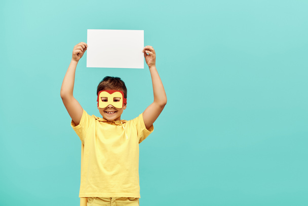 niño multirracial sonriente en traje de superhéroe amarillo con máscara sosteniendo papel en blanco sobre la cabeza sobre fondo azul, concepto del día internacional de los niños  - Foto, Imagen