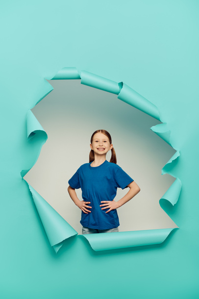 happy redhead preteen girl in t-shirt looking at camera while posing with hands on hips around blue torn paper hole on white background, International Child Protection Day concept  - Photo, Image