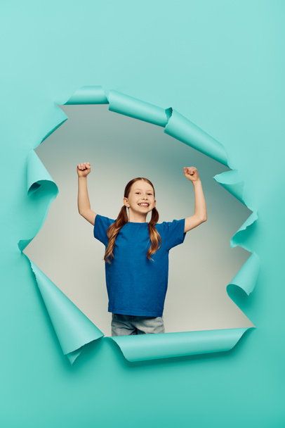 excited and redhead preteen girl in t-shirt looking at camera while showing power gesture around blue torn paper hole on white background, International Child Protection Day concept  - Foto, imagen