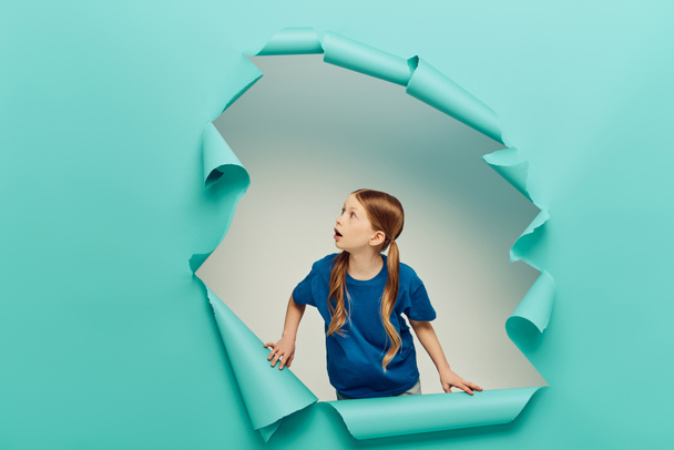 shocked redhead preteen girl in t-shirt looking at blue torn paper creating hole on white background, International Child Protection Day concept  - Photo, Image