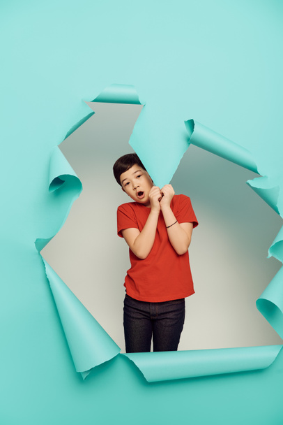 Shocked asian preteen boy red t-shirt looking at camera and touching hole in blue paper while celebrating child protection day on white background - Photo, Image