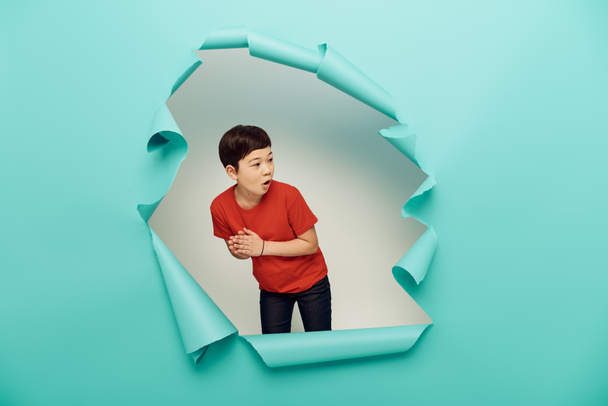 Surprised asian preteen boy in casual red t-shirt looking away during child protection day celebration behind hole in blue paper on white background - Foto, Imagen