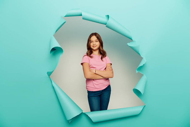 Smiling preteen girl in casual clothes crossing arms and looking at camera during child protection day celebration behind hole in blue paper background - Photo, Image