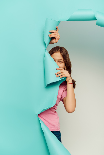 Preteen child in casual clothes cover face and looking in camera through hole in blue paper while hidden on grey background, International Child Protection Day concept  - Foto, Bild
