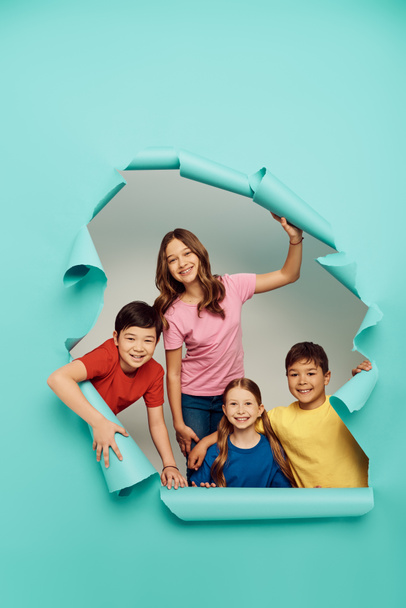 Cheerful group of multiethnic kids in colorful clothes looking at camera during child protection day while standing behind hole in blue paper background - Zdjęcie, obraz