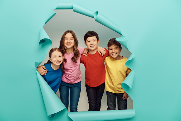 Smiling multiethnic preteen kids in colorful t-shirts hugging during children protection day celebration behind hole in blue paper background - Photo, Image