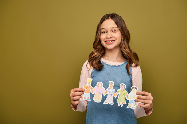 glimlachend tiener meisje in denim sundress houden getekend papier personages en kijken naar de camera tijdens de bescherming van het kind dag viering op kaki achtergrond - Foto, afbeelding