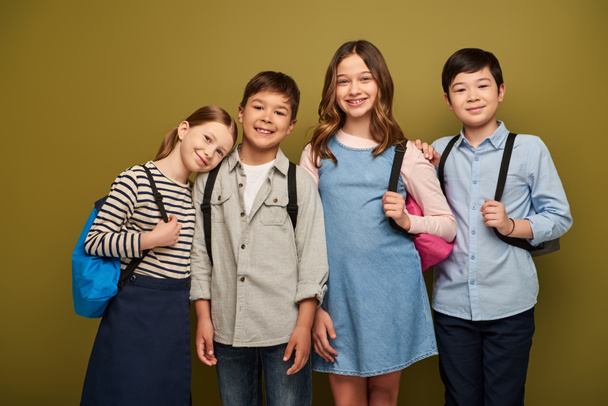 Позитивные межрасовые и подростковые дети в повседневной одежде, держащие рюкзаки и смотрящие в камеру во время празднования Дня защиты детей на фоне хаки - Фото, изображение